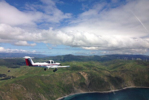 Piper Tomahawk ZK-TAW flies off the Mākara coastline