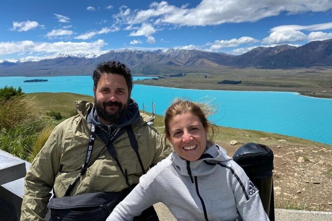 Couple on their accessible honeymoon in New Zealand, smiling beside their car with scenic mountain views in the background.