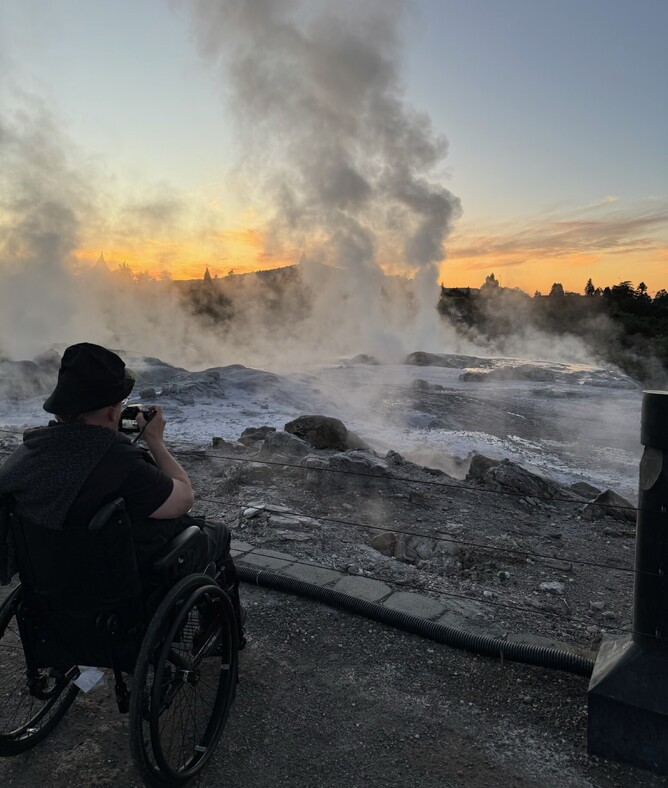 Man in wheelchair photographing an active geyser in Rotorua’s geothermal park