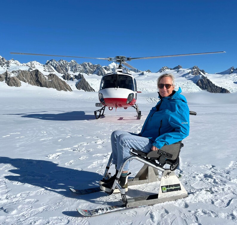 Woman in a snow-adapted wheelchair beside a helicopter in a snowy alpine landscape