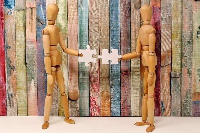 Two wooden figures working together to fit puzzle pieces, symbolising leadership collaboration and problem-solving