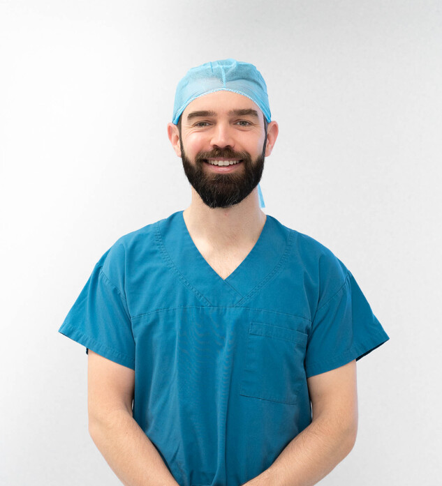 Hamish McLaren n an operating room, wearing surgical scrubs and a cap, operating a robotic surgical device while another medical professional works in the background.