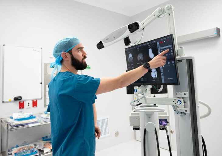 Hamish McLaren n an operating room, wearing surgical scrubs and a cap, operating a robotic surgical device while another medical professional works in the background.