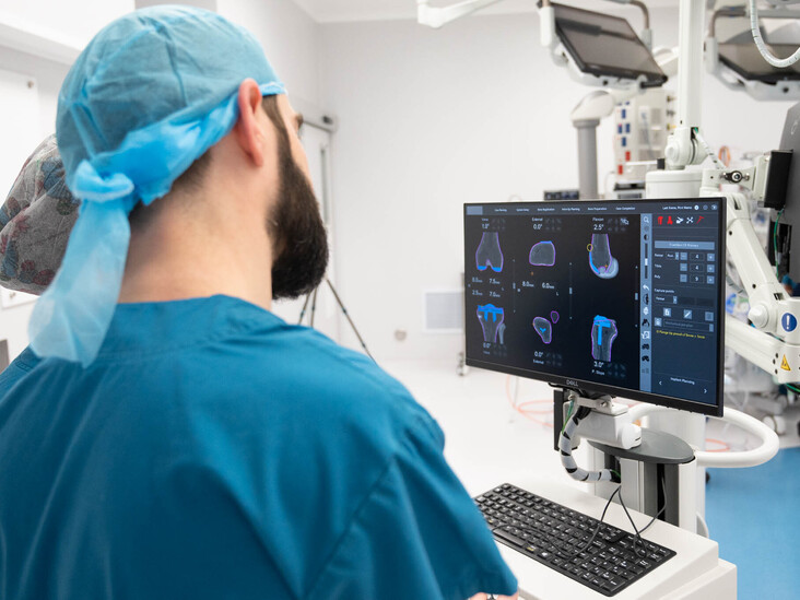 Hamish McLaren n an operating room, wearing surgical scrubs and a cap, operating a robotic surgical device while another medical professional works in the background.