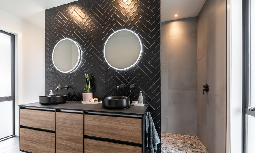 Modern black bathroom featuring a floating wall, black herringbone tile, with circular lit mirrors, and a walk-in pebble shower.