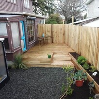 A tidy backyard space featuring a newly built timber deck and vertical timber fence. The deck includes a small step and raised garden beds on the right side filled with plants and vegetables. A gravel area borders the deck with potted plants and garden greenery, creating a neat, low-maintenance outdoor space.
