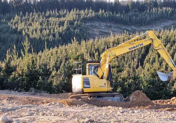A Komatsu PC138US excavator operating in a hilly, rural landscape, surrounded by young pine trees and native shrubs, with distant farmland visible in the background.