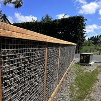 A close-up side view of a modern gabion-style fence made with timber framing and metal mesh panels filled with rocks. The fence lines a gravel driveway on a sunny day with a bright blue sky and green vegetation nearby.
