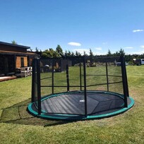 In-ground trampoline installation in a well-maintained backyard. Surrounded by a safety net, the trampoline sits flush with the grass surface. A modern house and utility vehicles are visible in the background, suggesting recent landscaping or installation work.
