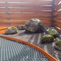 A modern timber slat fence surrounds a minimalist rock garden with large moss-covered boulders arranged on a bed of soil. In the foreground, a plastic honeycomb grid awaits ground cover installation, framed by timber edging.