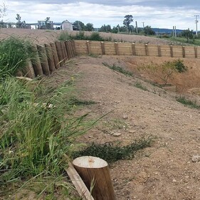 A landscaped rural backyard featuring a series of terraced retaining walls built from timber, designed to manage a steep slope. At the bottom, there’s a black modern-style house with outdoor furniture and a trampoline in the yard. The area is surrounded by open fields and greenery.