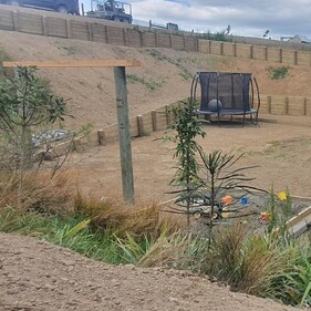 Landscaped backyard under development with timber retaining walls, a trampoline, rotary clothesline with laundry, a black house, and newly built wooden stairs leading up a hillside.