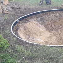 A large circular hole is dug in a grassy backyard, surrounded by a metal retaining ring, likely as a foundation for an in-ground trampoline installation.
