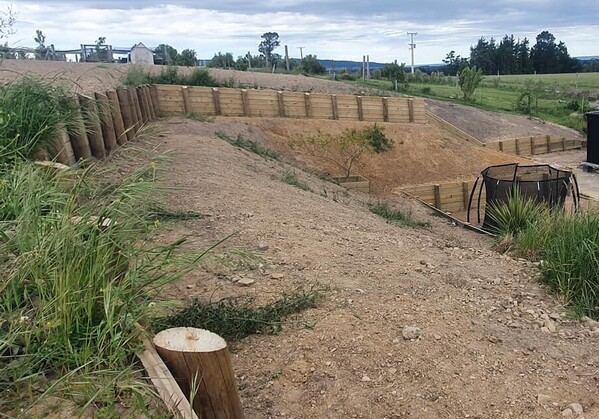 A landscaped rural backyard featuring a series of terraced retaining walls built from timber, designed to manage a steep slope. At the bottom, there’s a black modern-style house with outdoor furniture and a trampoline in the yard. The area is surrounded by open fields and greenery.