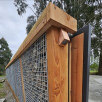 Close-up of a modern gabion fence with a sliding gate, featuring a timber frame and wire mesh panels filled with stones. The timber structure includes a small angled light fixture at the top, and the gate runs along a concrete track. Gravel ground surrounds the fence, and trees are visible in the background.
