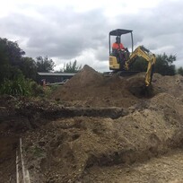 A worker operates a small excavator perched on top of a dirt mound at a construction site, moving earth for what appears to be early-stage site leveling or retaining wall preparation.