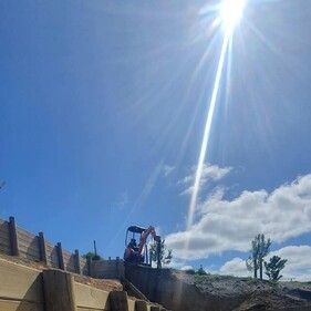 A bright, sunny day on a construction site featuring a timber retaining wall under construction. In the foreground, long timber planks and vertical support posts are laid out. At the top of the slope, an excavator is drilling or compacting, silhouetted by the sun overhead casting a strong sunbeam through the clear sky.