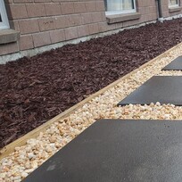 Straight pebble pathway bordered with dark square pavers, running alongside a house. The path is flanked by a mulched garden bed on one side and freshly tilled soil on the other, leading toward a wooden deck in the background.
