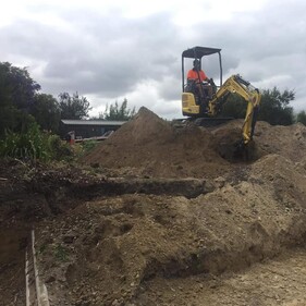 A worker in a bright orange shirt operates a small yellow excavator on top of a large mound of soil at a residential construction or landscaping site, with cloudy skies overhead and surrounding greenery.
