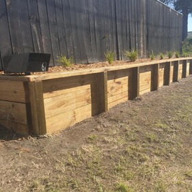 Freshly built timber retaining wall with vertical posts and horizontal planks, topped with mulch and small decorative plants, running along a black wooden fence beside a suburban driveway.