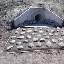 Concrete culvert outlet surrounded by compacted gravel and framed with a rectangular rock bed for erosion control. The rocks are neatly spaced on a sloped surface for effective water dispersion.