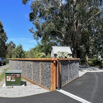 A modern gabion-style fence constructed with metal mesh panels filled with rocks, framed by timber posts and caps. The fence lines a freshly paved driveway under a bright blue sky, with tall trees and a small corrugated shed in the background.