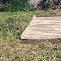 A small timber platform or low bridge built over grassy terrain, bordered with soil at the base and framed by simple timber edging.