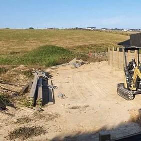 A construction site with a small yellow excavator parked on sandy ground, in front of a partially completed wooden retaining wall supported by timber posts. The area is surrounded by open fields and bordered by a tall black fence.