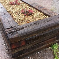 Rustic raised garden bed constructed with weathered railway sleepers, filled with mulch and planted with small flowering shrubs. The structure sits beside a paved area with signs of rainwater runoff.
