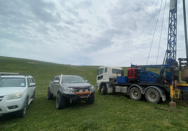 Field drilling setup with a large blue and white rig truck in operation, flanked by two support vehicles on green open farmland under a cloudy sky.