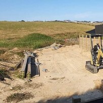  construction site showing early progress on a timber retaining wall. A small yellow digger is stationed in front of the freshly installed posts and horizontal timber planks, ready for further earthmoving.