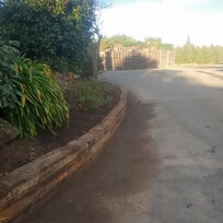 Freshly edged and tidied roadside garden with curved stone retaining border, featuring dense green foliage and neatly turned soil. A clean asphalt surface runs alongside, with wooden pallets stacked in the background.