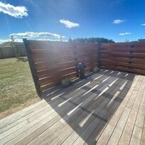 A modern outdoor patio area with a horizontal timber privacy screen and wooden decking. The fence casts striking shadows on the deck under bright sunlight. The space includes a small decorative water feature and several rocks along the fence base.
