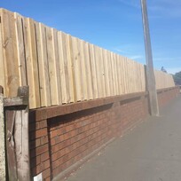 Newly installed vertical timber fence atop an existing red brick boundary wall along a suburban sidewalk. A concrete power pole intersects the fence line, and cars are parked along the roadside under a clear blue sky.