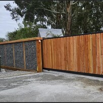 A modern automated sliding gate made with vertical timber slats framed in black steel, integrated seamlessly with a gabion rock wall and timber posts. 
