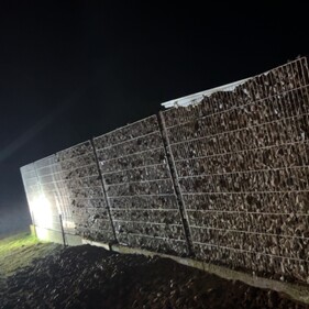 A night-time view of a partially filled gabion retaining wall made from metal mesh and filled with rocks. A bright construction light illuminates the scene from the left side, casting shadows on the rocky ground. The text “She’s filling up” is overlaid vertically on the wall.