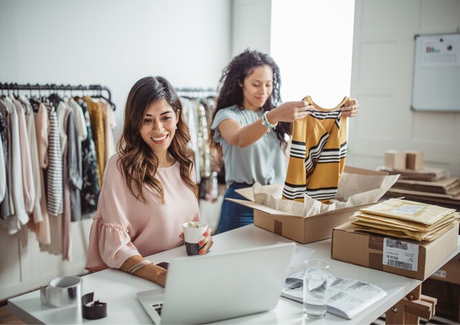 Two ladies packaging clothes in a small warehouse and working on the laptop
