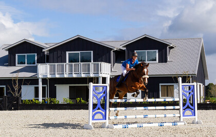 Horse and rider jumping in a free draining arena in front of a barn and stables