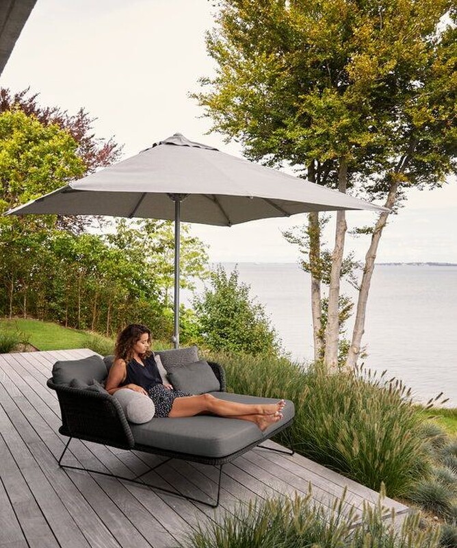 A woman is reclining on a daybed that sits on a timber deck beneath a large shade umbrella. The deck over looks the ocean and there are garssy plants around it, and a few large trees in the background.