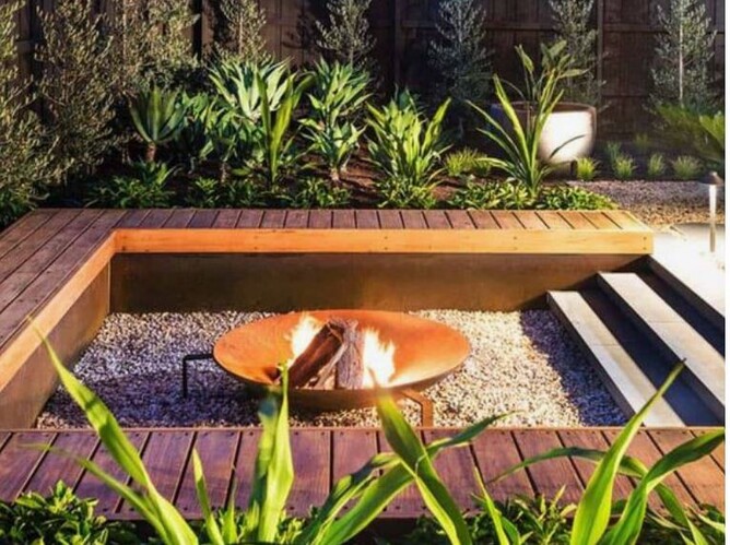 A backyard landscaping idea showing a sunken firepit area with built in timber seats surrounding a sunken fire pit area. /in the centre of the garden space sits a rust-look firebowl. The space has lush planting in the background.