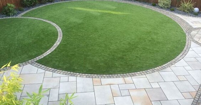 A lovely lawn area in a backyard featuring to circles of lush green grass intersected by a band of small square pavers. The same pavers wrap the periphery of both the lawn s and match the larger, autumn-toned pavers of the adjacent patio area.