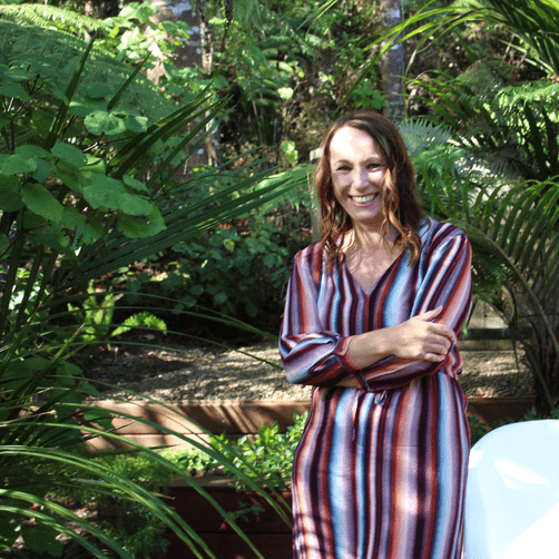 A woman wearing a blue and red striped dress striped dress. She has shouder length brown hair. Her arms are crossed and shes smiling.
