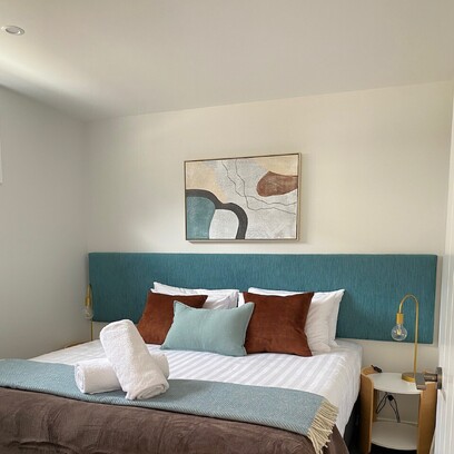 A light walled modern bedroom with an artwork on the wall above a large double bed, teal headboard, throw cushions, and beside table with lamp.