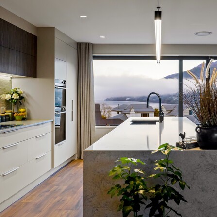 Looking out over a designer kitchen with light stone bench tops, bar pendant lighting, wooden floors and a window view out to the lake.