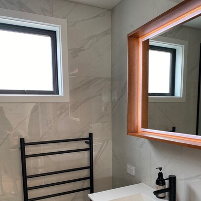 A marble look tiled bathroom with a white topped vanity, black heated towel rail, and framed mirror.