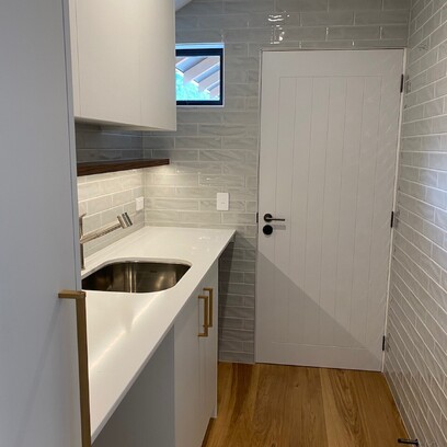 A fully functional laundry with light grey tiled walls, white bench top and white cabinetry with brass handles.