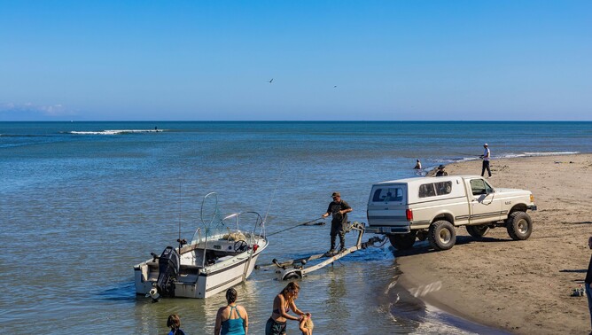 White 4WD vehicle backing a small boat trailer into the water at a beach boat ramp, with people standing nearby enjoying the sunny day and calm sea.