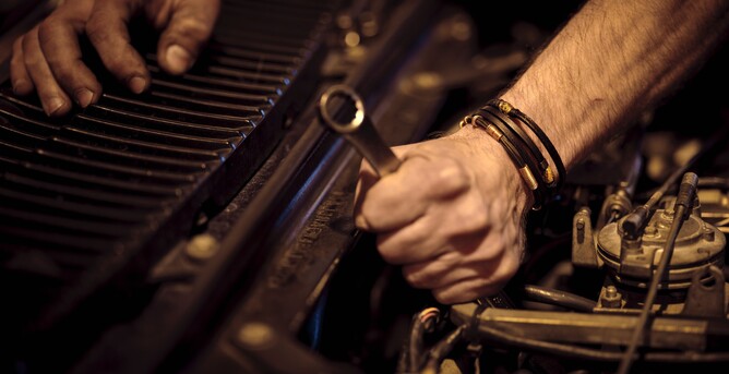 Mechanic working on a car engine, gripping a wrench with one hand near the radiator and engine components in a dimly lit workshop.