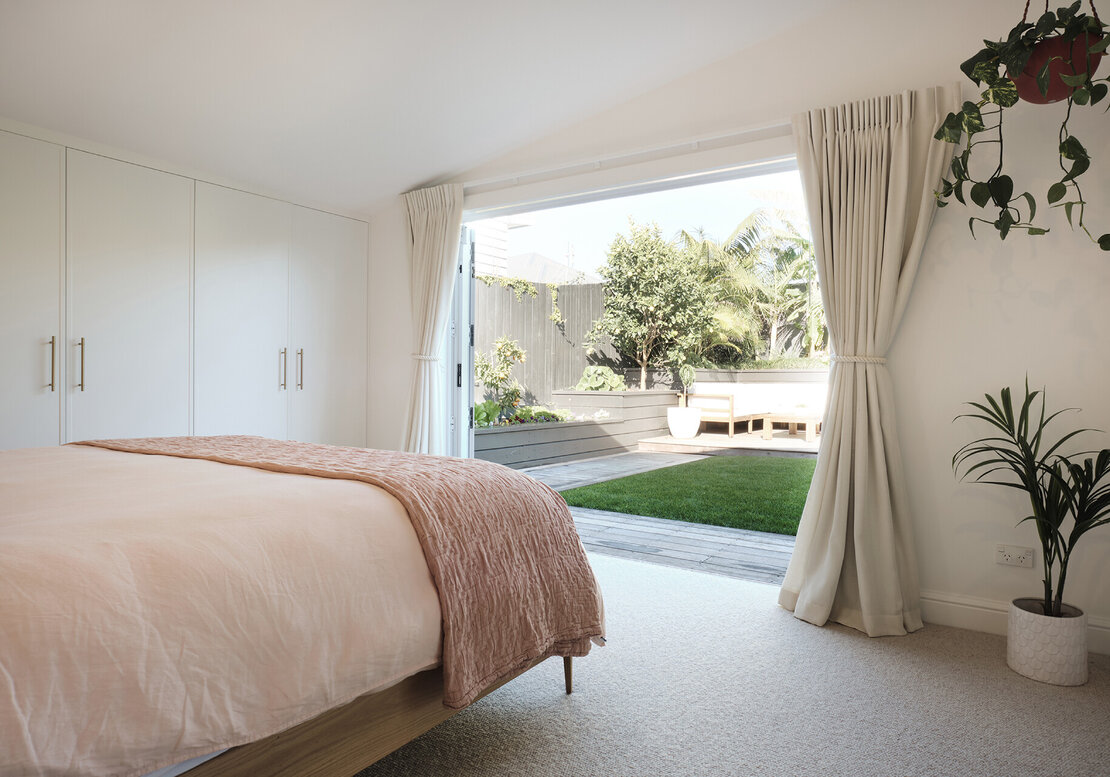 A newly renovated bedroom with new wardrobe doors, carpet, walls, curtains, bed, and bifold patio doors. The patio doors are open, looking out to the garden.