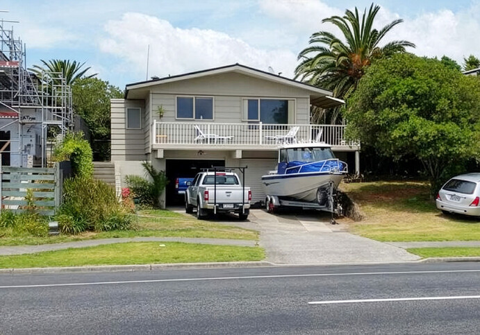 The image shows a home before a builder's renovation in Papamoa, Bay of Plenty. It's an old split-level beach bach with two single garages on the ground floor and living areas and a deck on the second level. Two cars and a boat are parked outside.